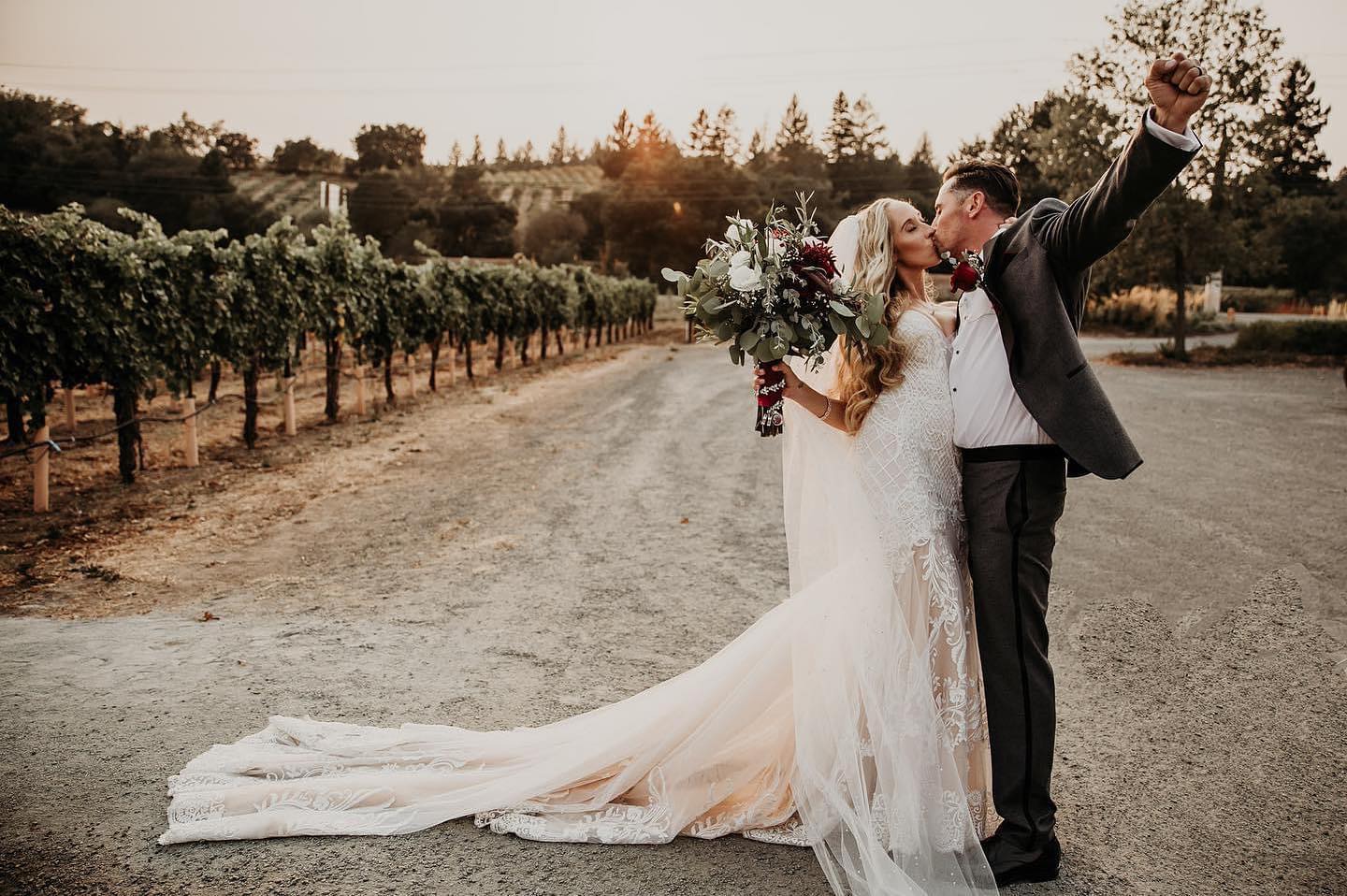 Bride and groom kissing holding bouquet on gravel patch with vineyards in background