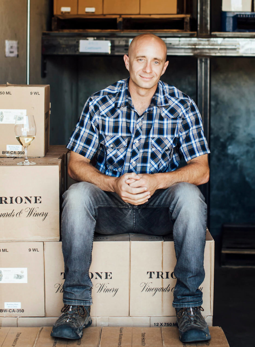 Man sitting on stacked wine boxes; glass of wine next to him.