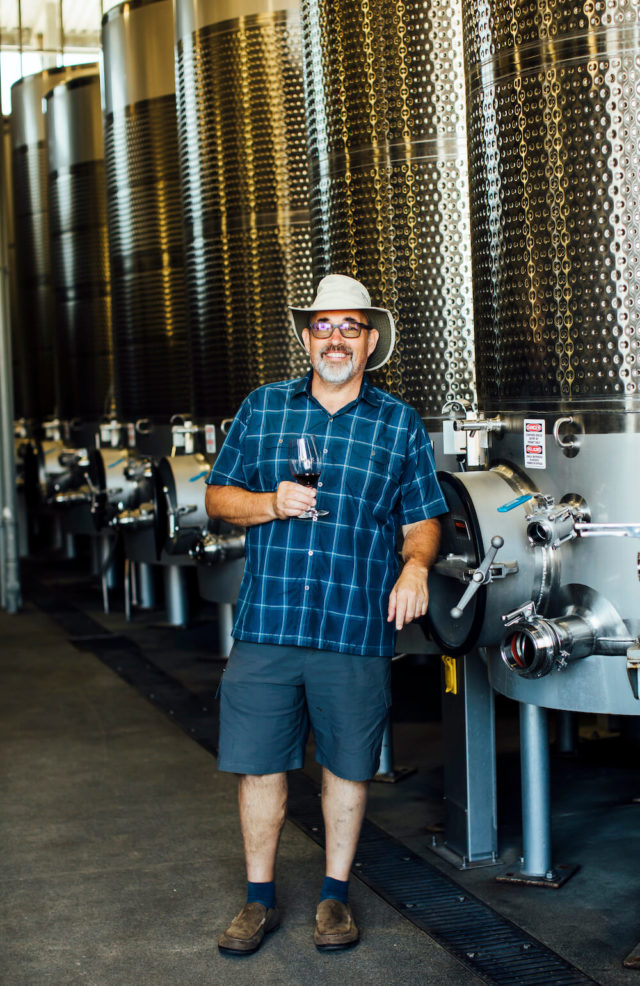 Man standing in front of metal tanks holding a glass of wine.